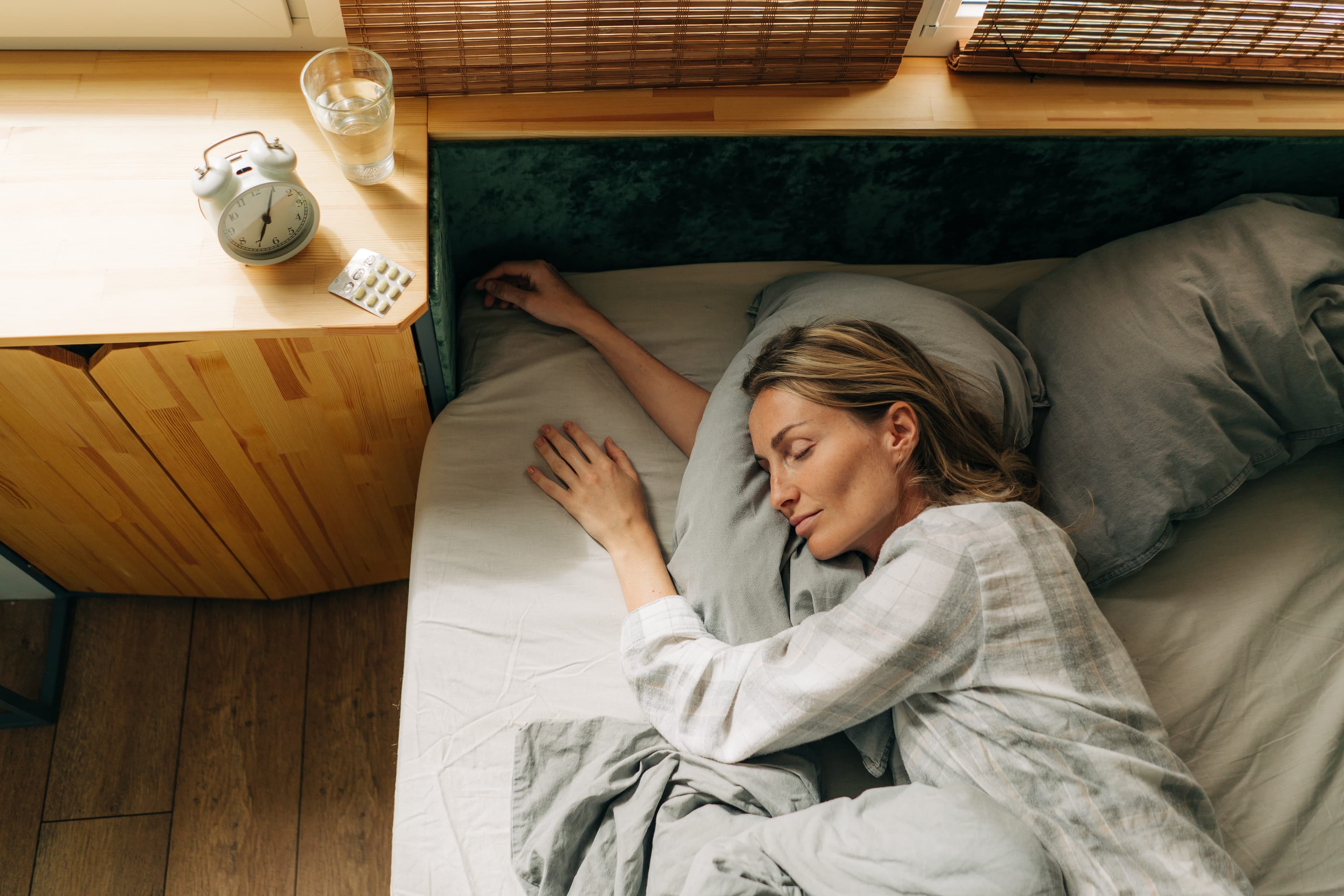 A high-angle view of a woman sleeping deeply in bed with a blister pack of pills and a glass of water on the bedside table, visually illustrating the risks of Zopiclone overdose and the importance of adhering to prescribed dosage limits.